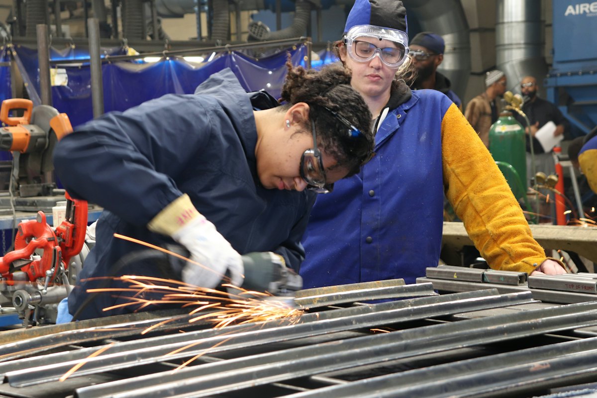 A learner cutting metal in a trade school workshop while an instructor watches